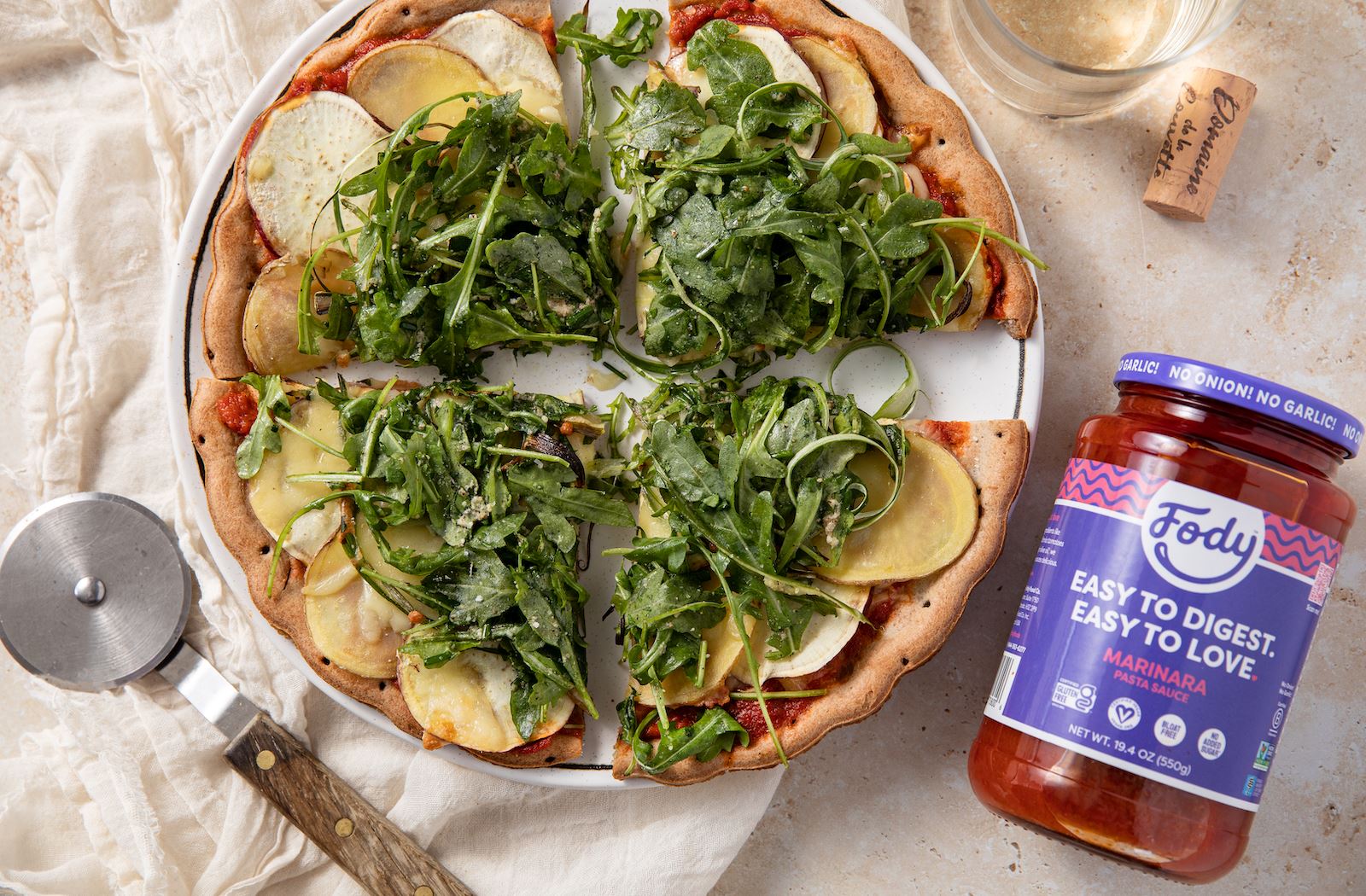 An image of Fody's classic veggie pizza on an elegant table setting beside a pizza slicer and a jar of marinara sauce. 